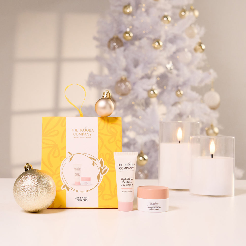 The Jojoba Company holiday gift set arranged on a white surface in front of a softly blurred white Christmas tree with gold ornaments. The set includes a yellow and white gift box, a pink and white lotion tube, a small pink-lidded cream jar, and two lit white candles in clear glass holders, with a gold ornament beside the box.