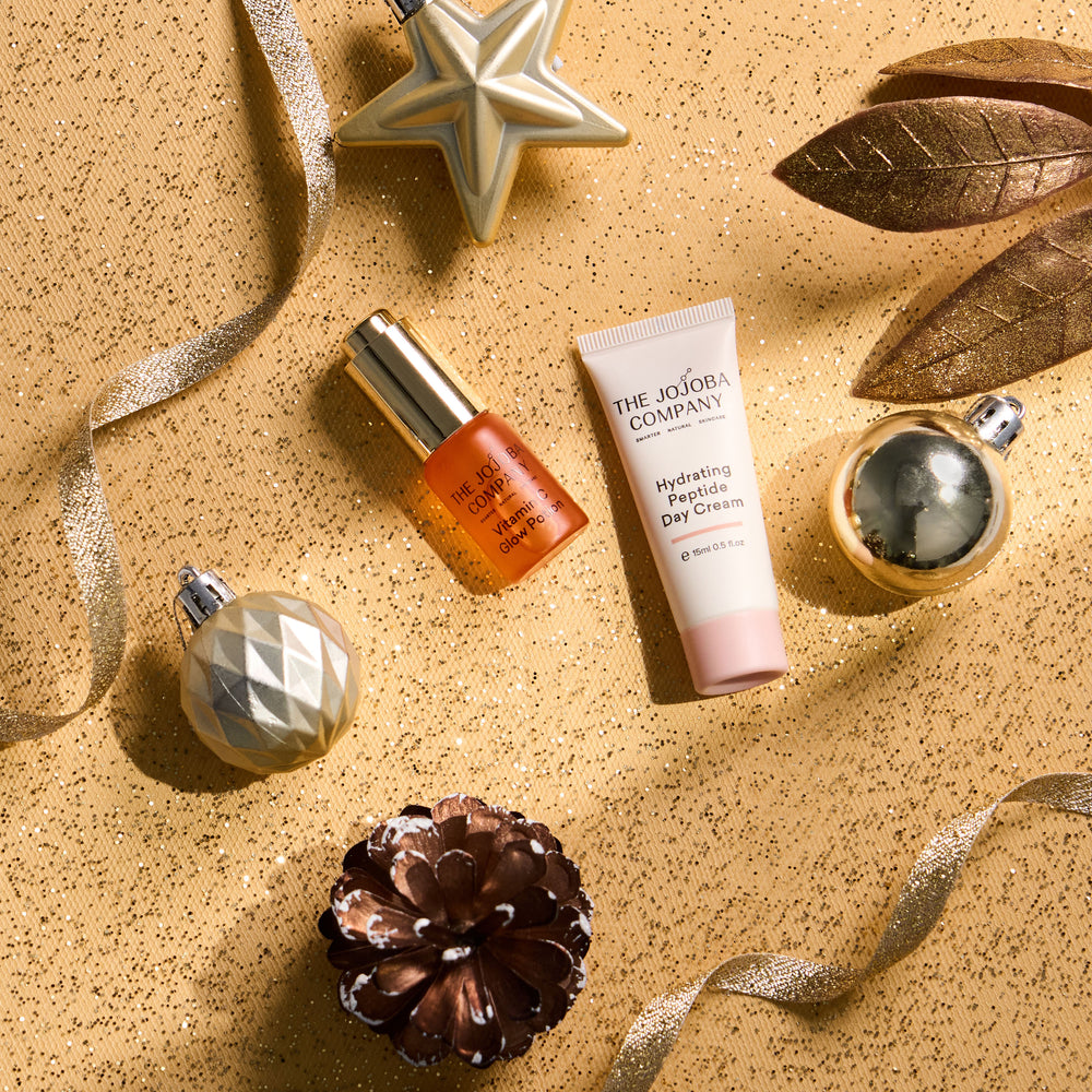 A styled flat-lay of two skincare products from The Jojoba Company on a glittery gold background: an orange-toned serum bottle with a gold cap and a white hydrating face cream tube with a pink base. Decorative holiday ornaments, ribbons, a star, and a pinecone surround the products.