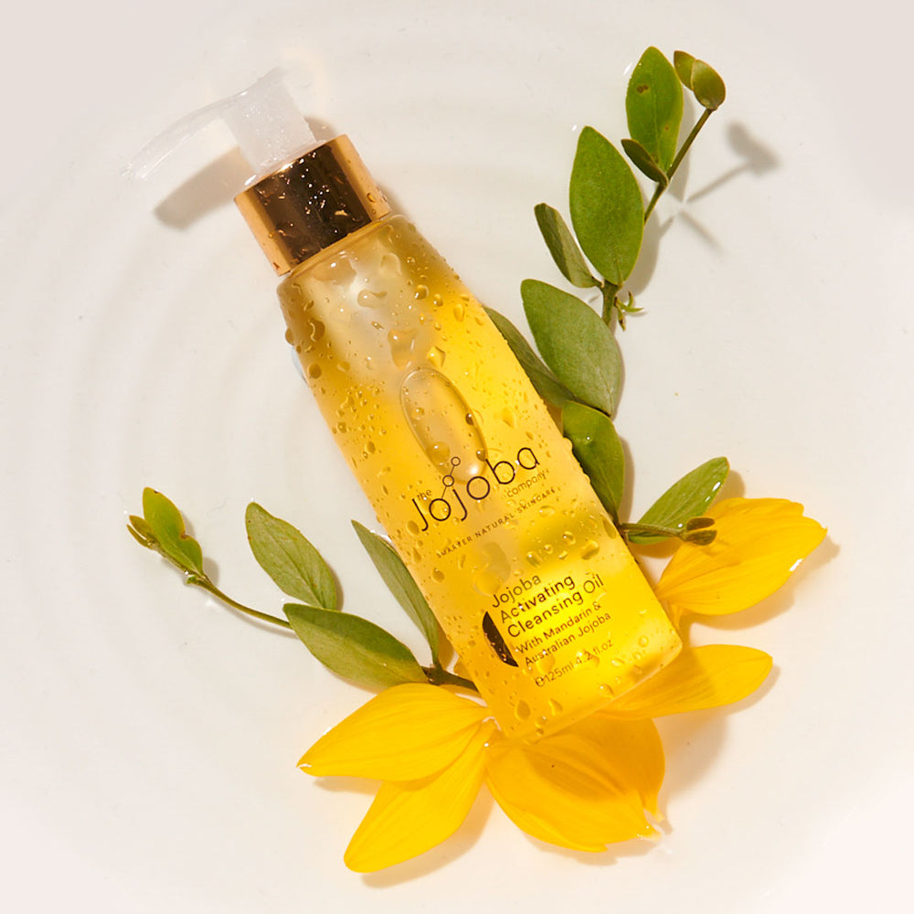 A bottle of jojoba cleansing oil with droplets on it, surrounded by yellow petals and green leaves.