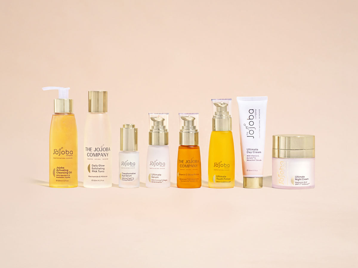 A range of The Jojoba Company skincare products against a beige background.