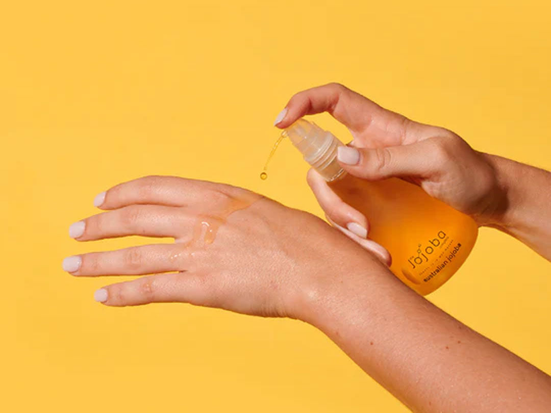 Person applying oil to hand from dropper bottle against yellow background.