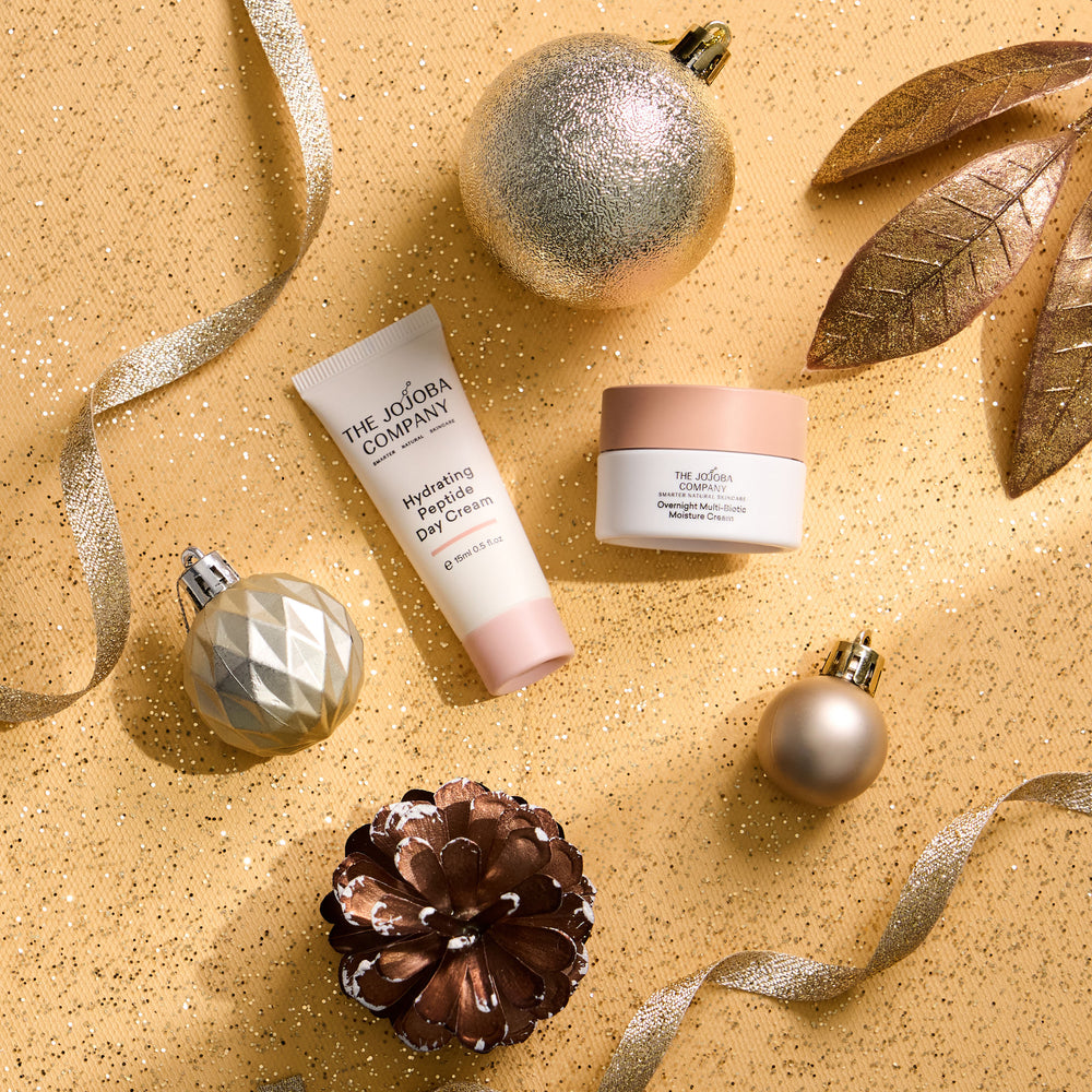 Two skincare products from The Jojoba Company arranged on a glittery gold background with holiday ornaments, ribbons, and leaves. The products are a Hydrating Hand Cream in a white tube with a pink cap, and a Hydrating Face Cream in a white jar with a pink lid.