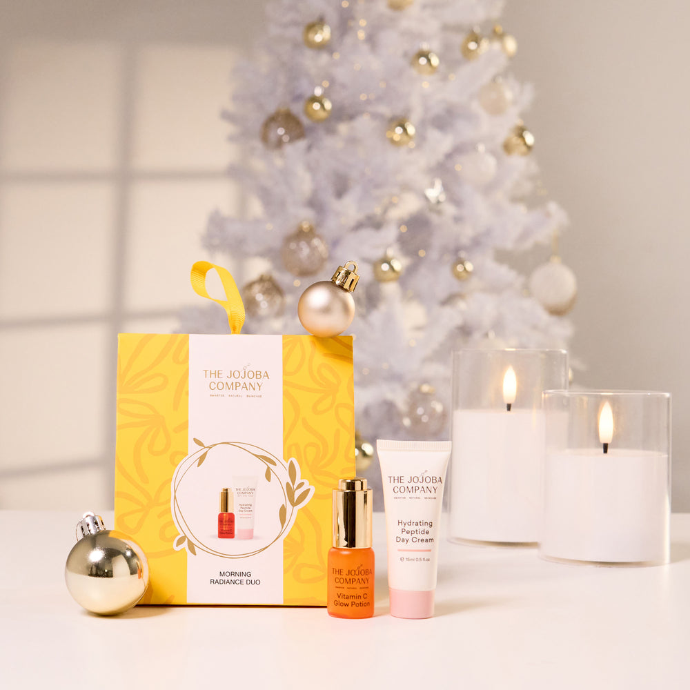 The Jojoba Company Morning Enthusiasm Day gift set displayed on a white surface in front of a decorated white Christmas tree. The set includes a yellow and white gift box, an orange face oil bottle with a gold cap, a pink face cream tube, and a gold ornament.