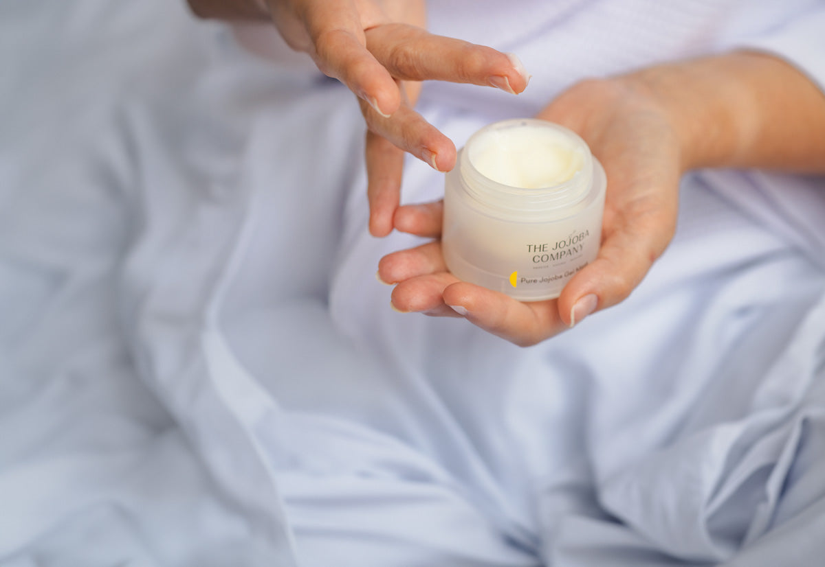 Person holding a jar of The Jojoba Company cream, preparing to apply skincare product.