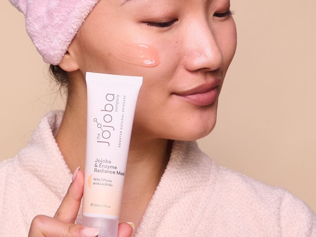 Person holding a Jojoba & Enzyme Radiance Mask tube, wearing a pink headband and white robe.