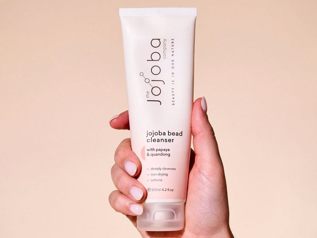 A hand holding a tube of jojoba bead cleanser against a beige background.