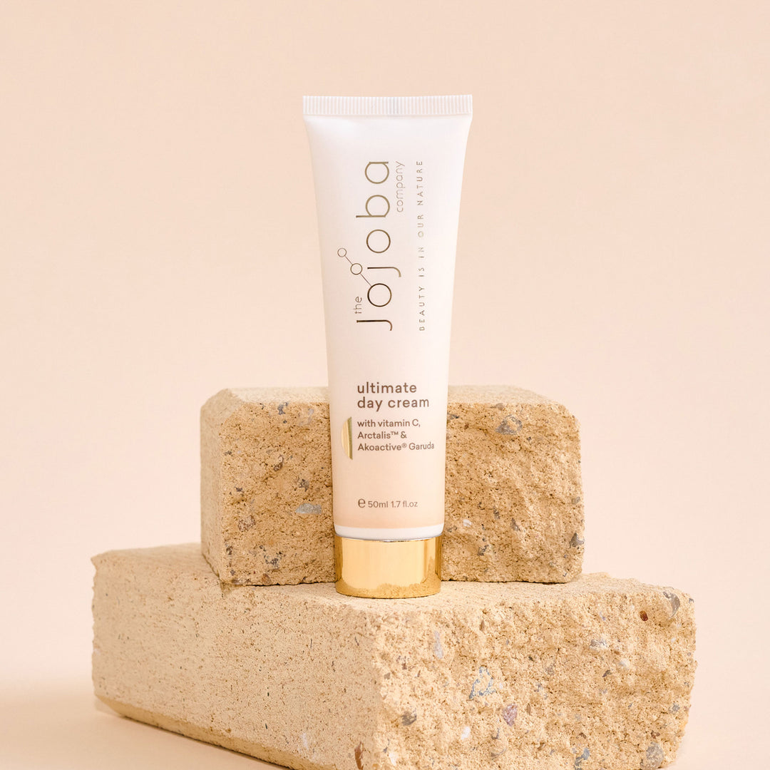 A tube of "The Jojoba Company" ultimate day cream on a beige brick, with a neutral background.