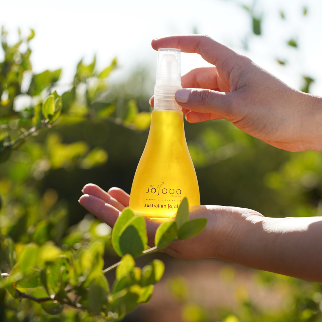 A hand holds a bottle of jojoba oil with greenery in the background.